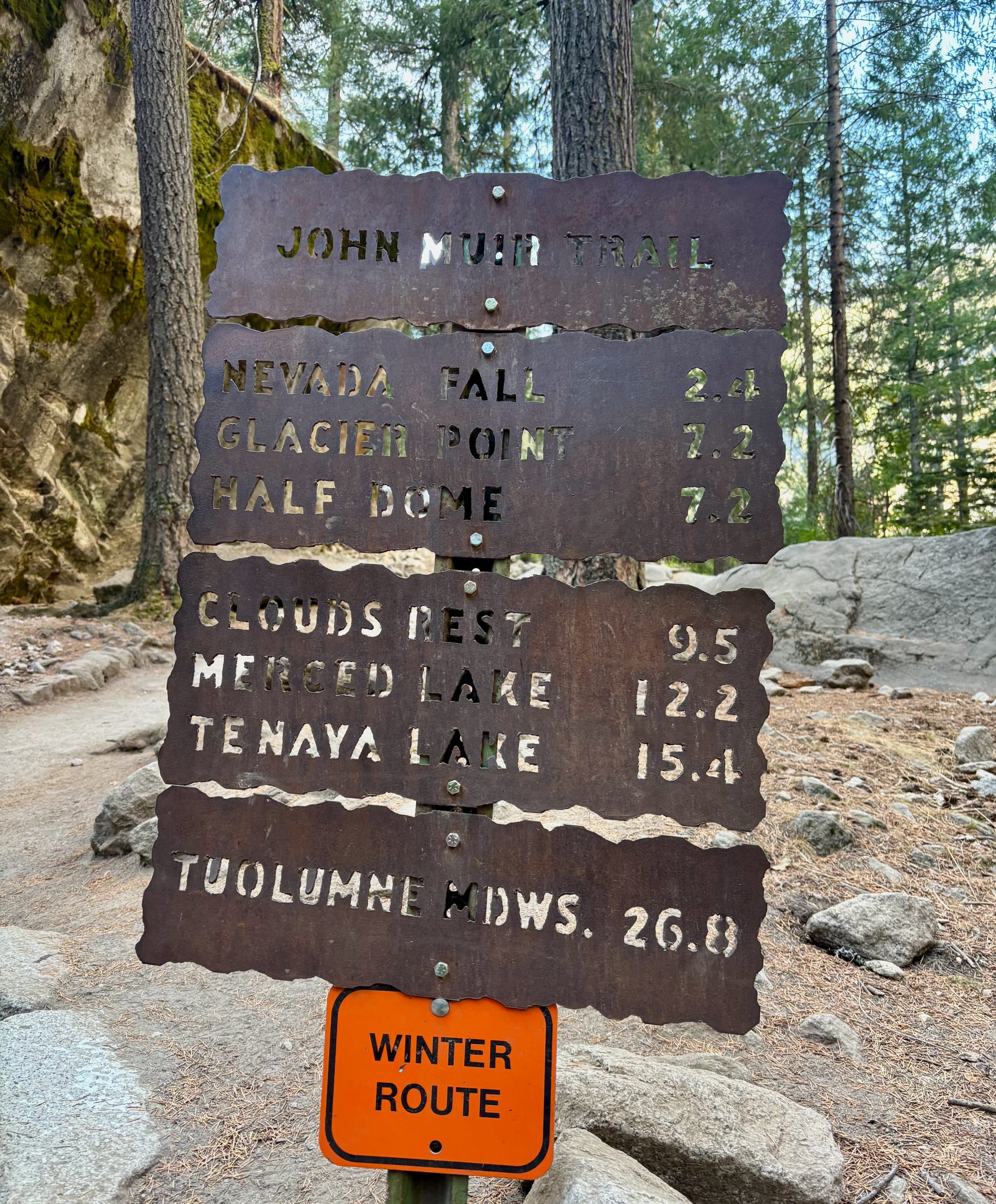 Trail sign in a forest indicating distances to various locations on the John Muir Trail, with a small "Winter Route" sign below.