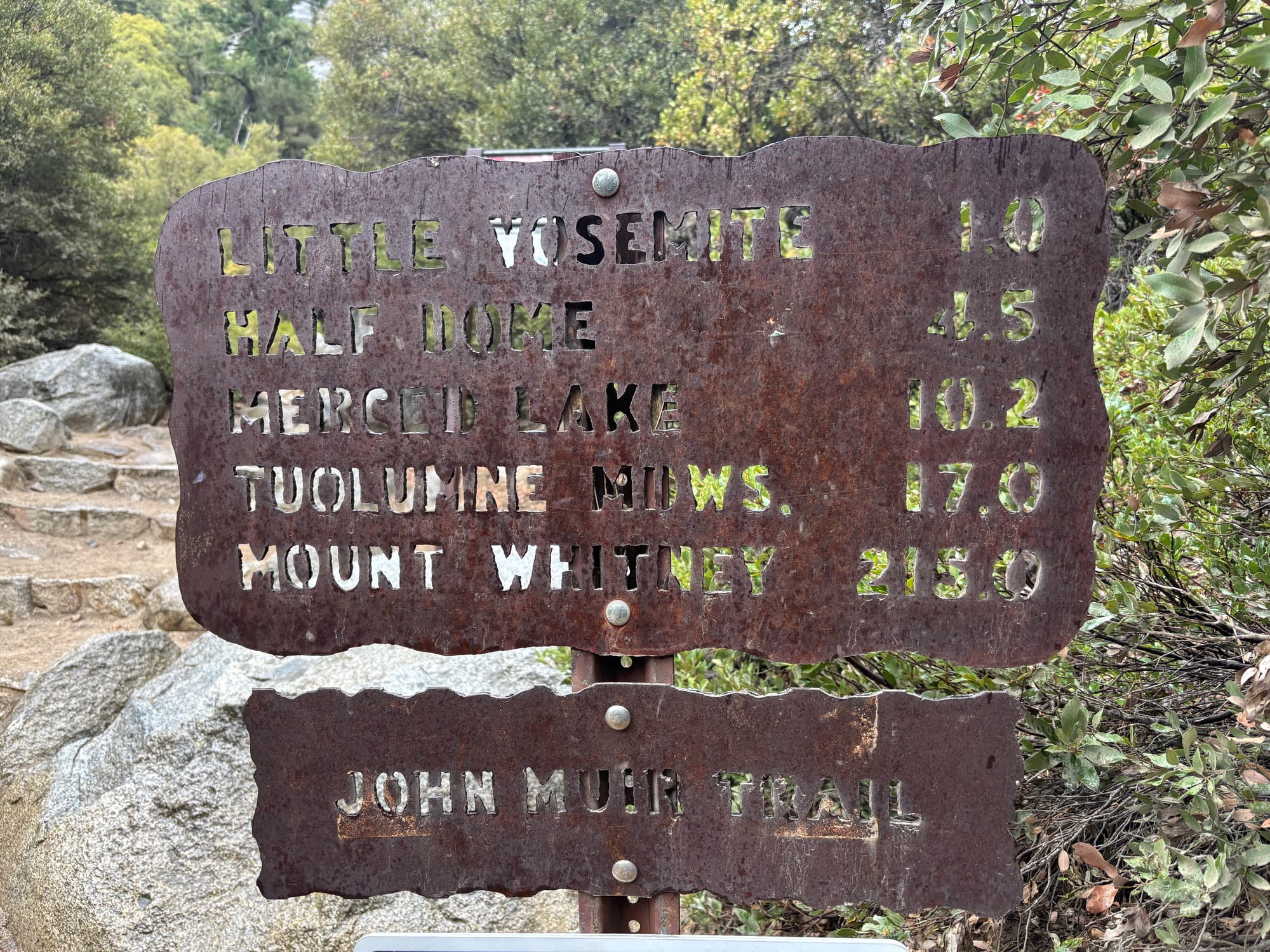 Rustic metal trail sign with distances to Little Yosemite, Half Dome, Merced Lake, Tuolumne Meadows, and Mount Whitney on the John Muir Trail.