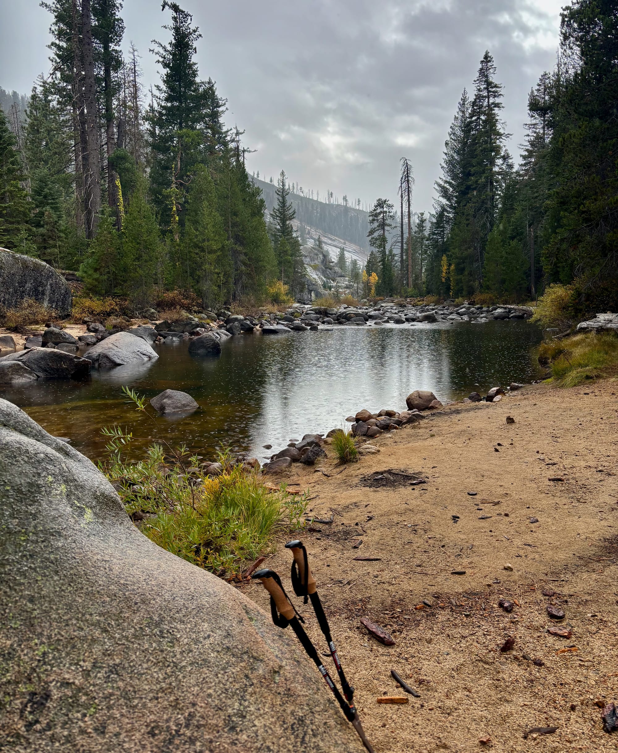 A serene river scene with rocks, surrounded by tall evergreen trees and a misty sky. A hiking trail is visible in the foreground, marked by a pair of trekking poles.