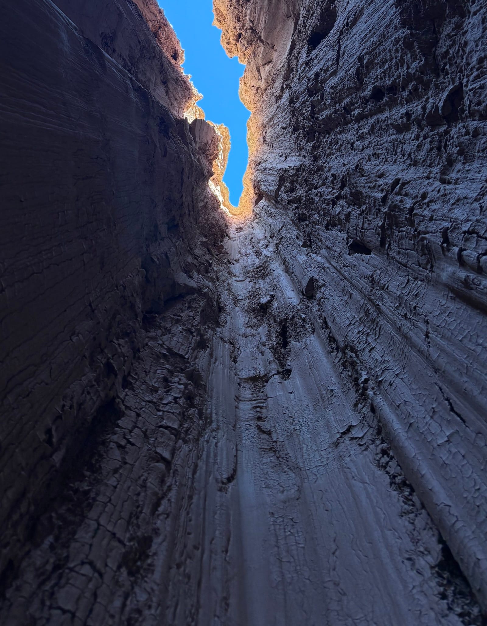 View upwards through a narrow slot canyon, showing vertical striations in the rock walls against a blue sky.