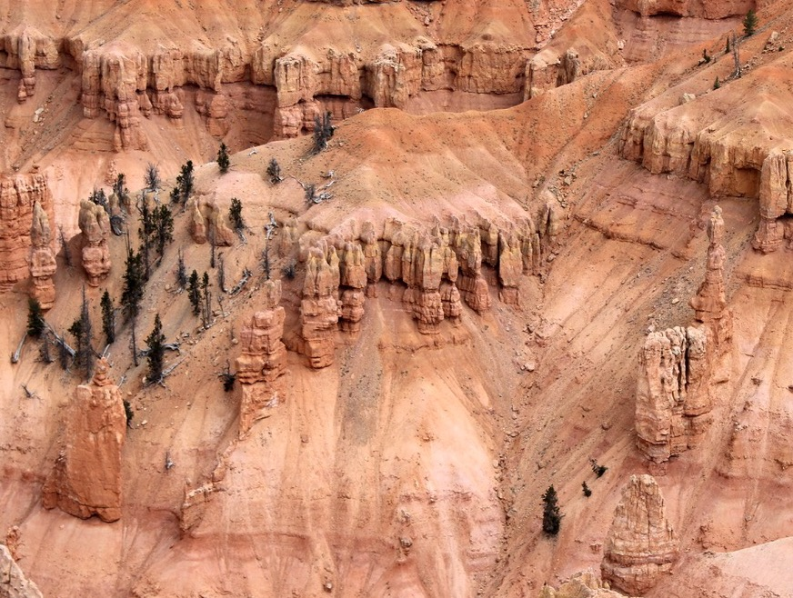 A close-up aerial view of a hoodoo landscape. Towering rock spires of varying shapes and sizes dominate the scene, showcasing a range of earthy tones including orange, red, pink, and cream