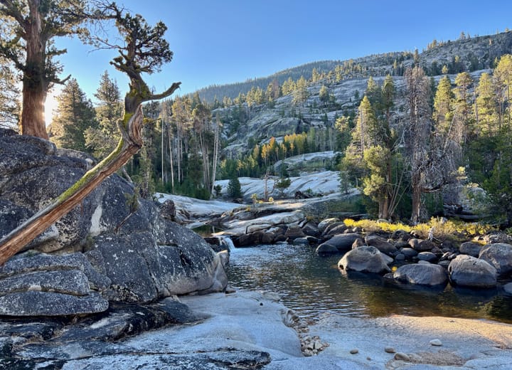 A serene mountain landscape with a rocky stream surrounded by trees and hills under a clear blue sky.