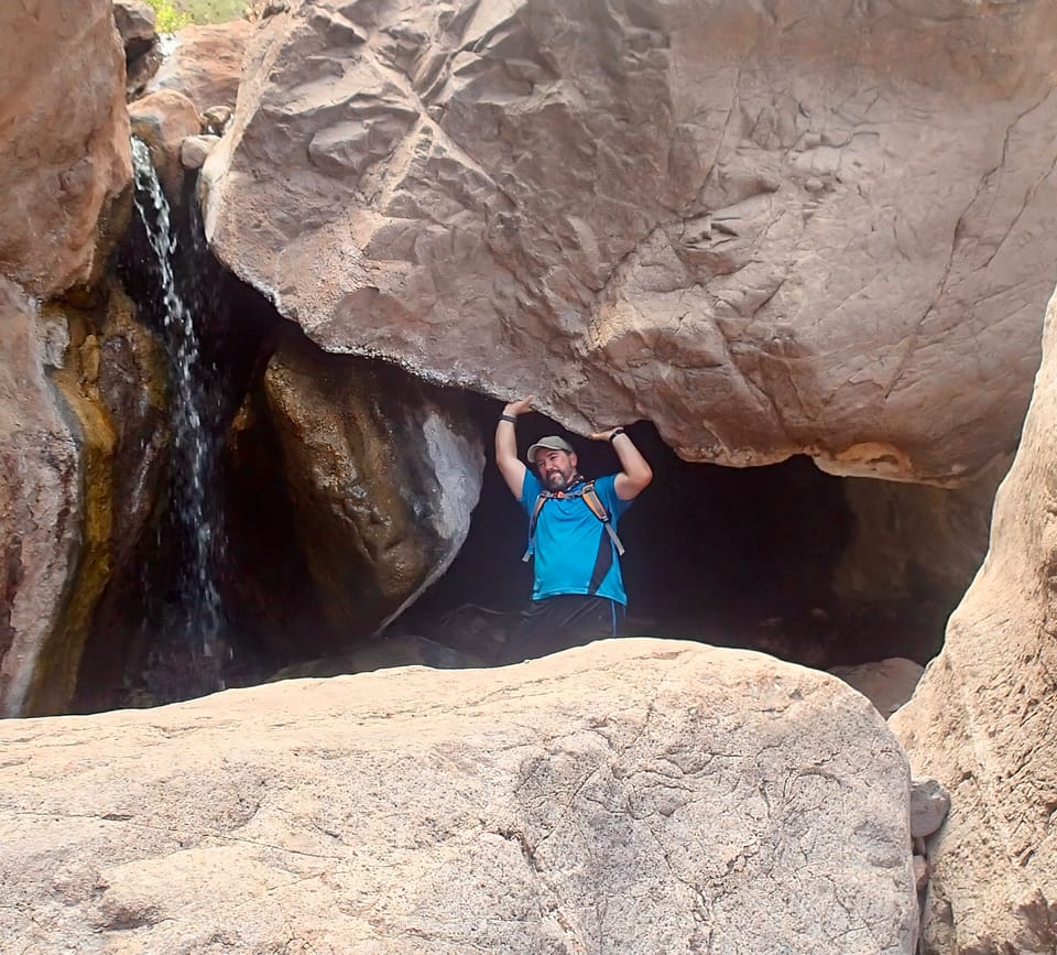 A person in a blue shirt is standing beneath a large rock formation, with a small waterfall nearby. 