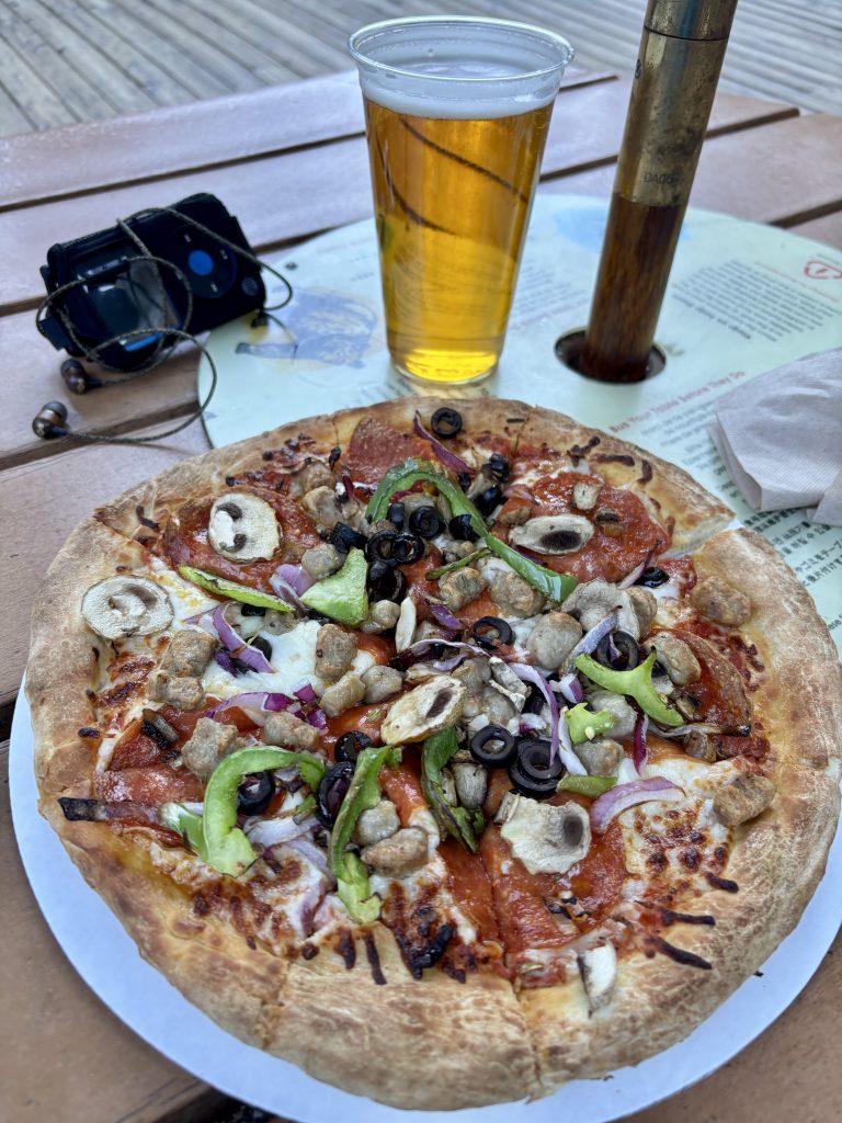 A pizza sits on a wooden table, covered in toppings, with a frosty beer and iPod beside it.