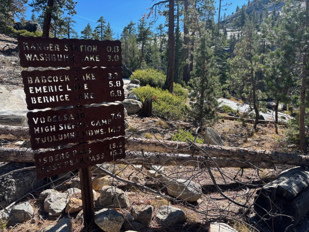 A weathered trail marker post displaying hiking distances to nearby lakes, set within a rocky and forested area.