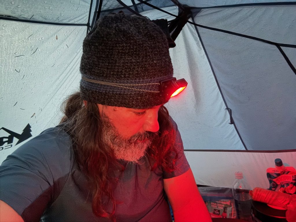 A close-up portrait of a person inside a tent. The person has long, dark hair and a full, graying beard. They are wearing a dark, knitted beanie and a headlamp casts a warm, reddish glow on their face. They appear to be looking down at something, possibly a map or device, illuminated by the headlamp. The tent walls are visible in the background, giving a sense of being in a sheltered, possibly camping environment.