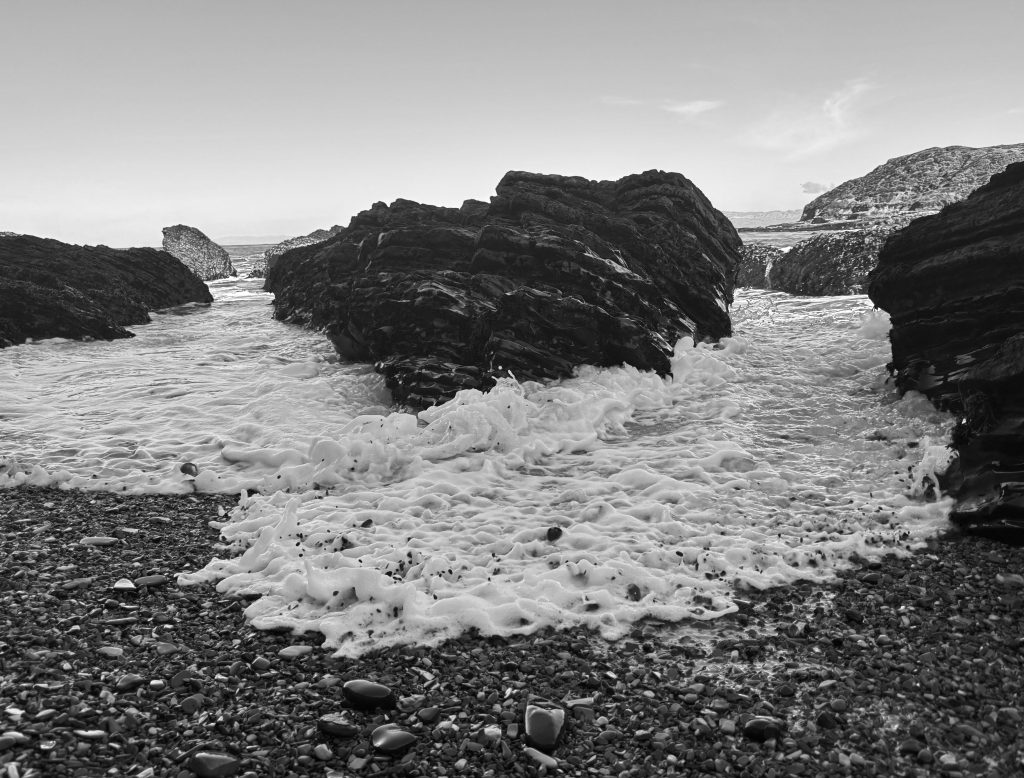 Black-and-white photo of foamy ocean waves pushing through rocky coastal crevices onto a pebble-strewn beach, with layered sea cliffs in the background.