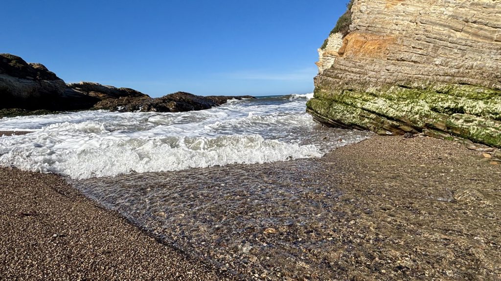 Small rocky beach with clear shallow waves washing toward the shore, flanked by layered sea cliffs with green algae under a bright blue sky.