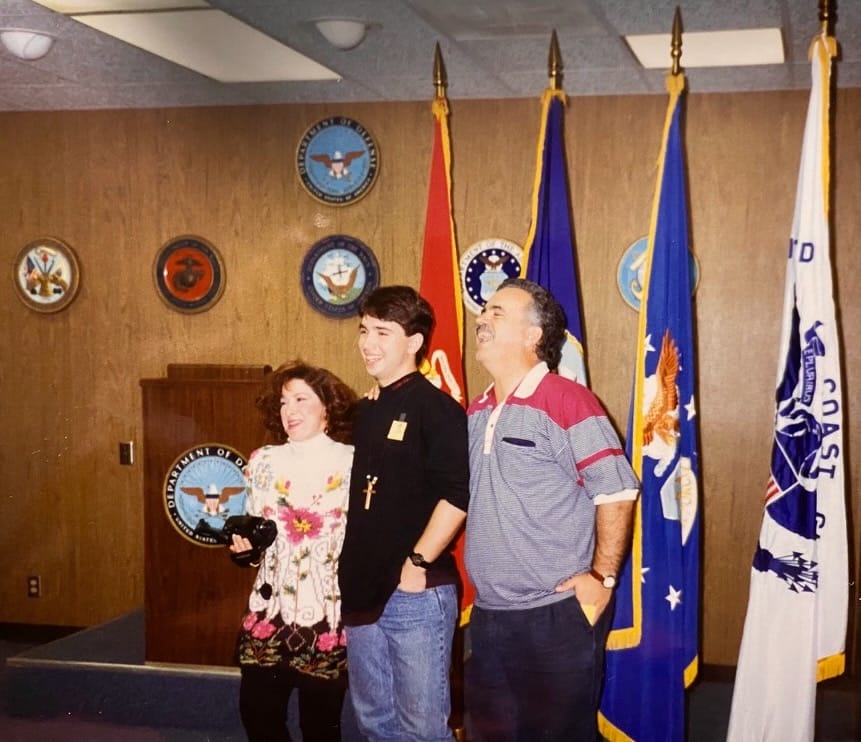 Three people stand smiling in front of military service flags and a Department of Defense podium. The young man in the center wears a black shirt and cross necklace, while the woman on the left holds a camera and wears a colorful floral outfit. The man on the right wears a striped polo shirt and has his hands in his pockets. The setting appears to be an indoor military or enlistment office.