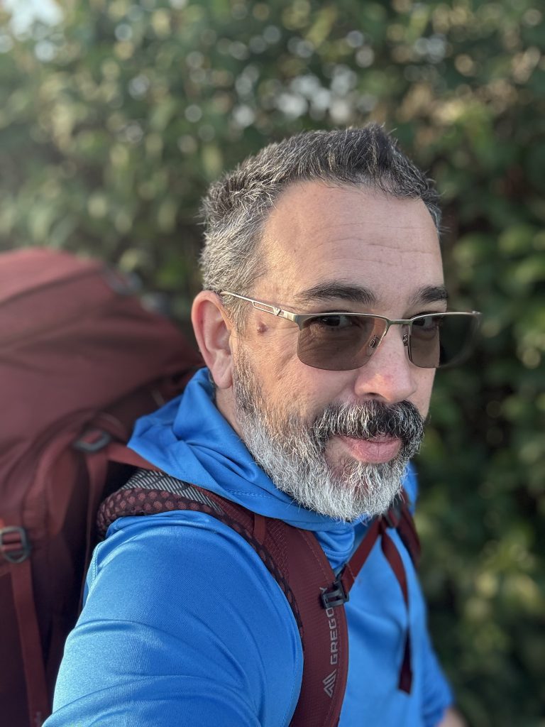 Middle-aged man with gray beard and sunglasses smiling while wearing a blue hoodie and a large red backpack outdoors.