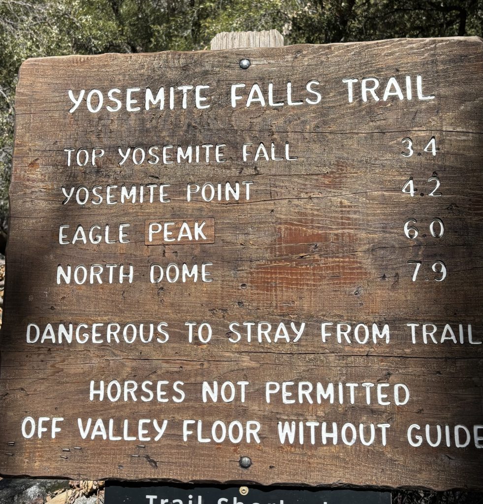 Wooden trail sign reading "YOSEMITE FALLS TRAIL" with distances: Top Yosemite Fall 3.4, Yosemite Point 4.2, Eagle Peak 6.0, North Dome 7.9. Warning lines: "DANGEROUS TO STRAY FROM TRAIL" and "HORSES NOT PERMITTED