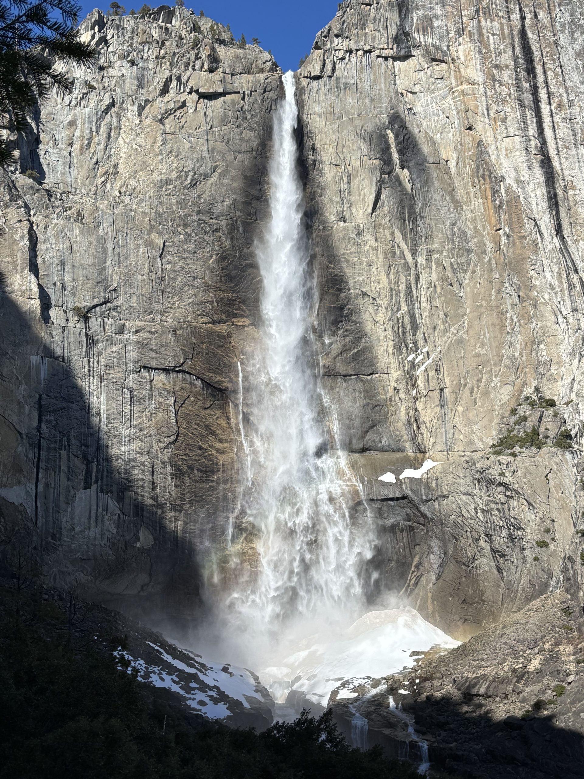 Tall waterfall cascading down a granite cliff with spray and a faint rainbow, framed by trees and rocky terrain.
