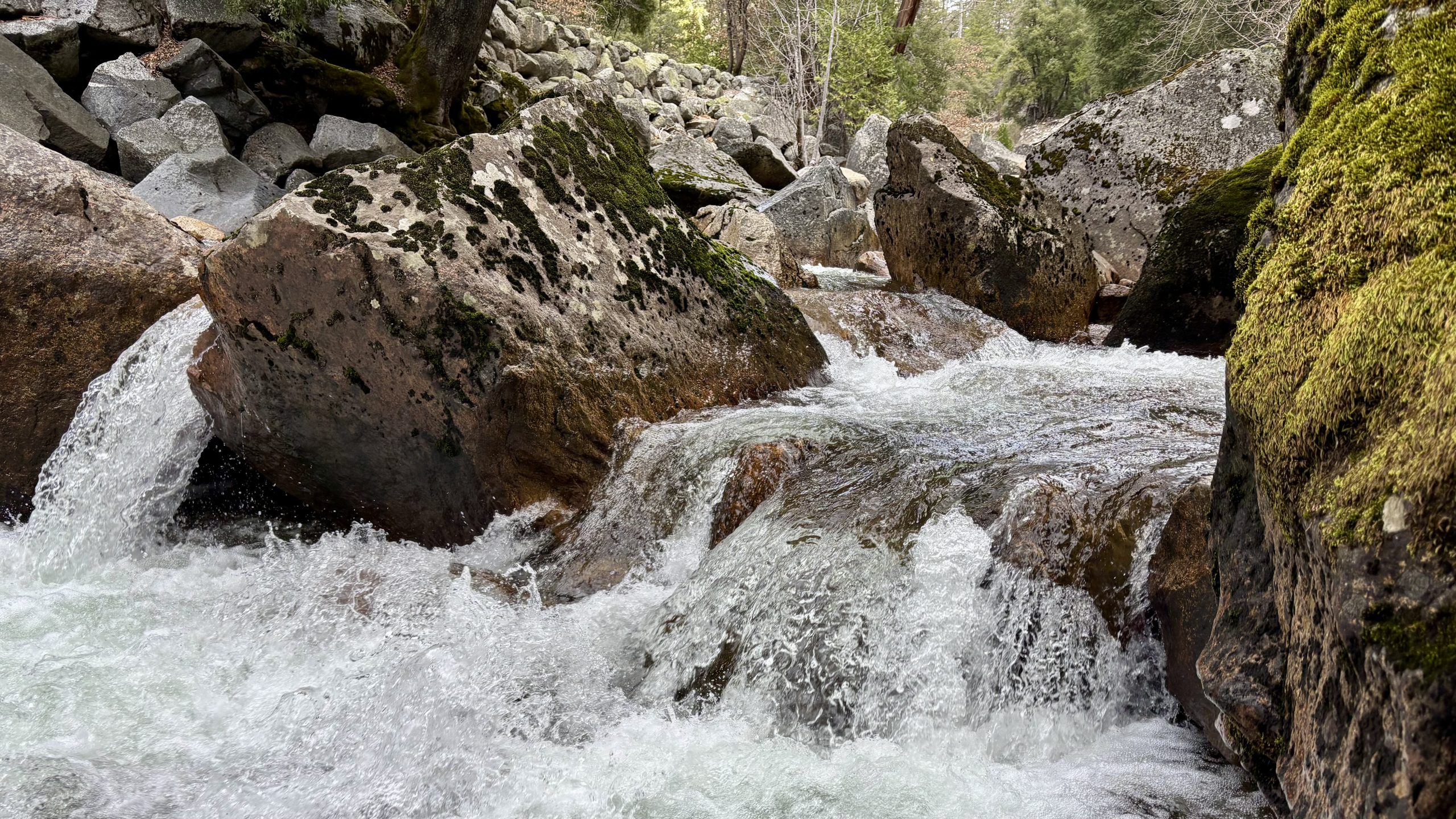 Tall waterfall cascading down a granite cliff with spray and a faint rainbow, framed by trees and rocky terrain.