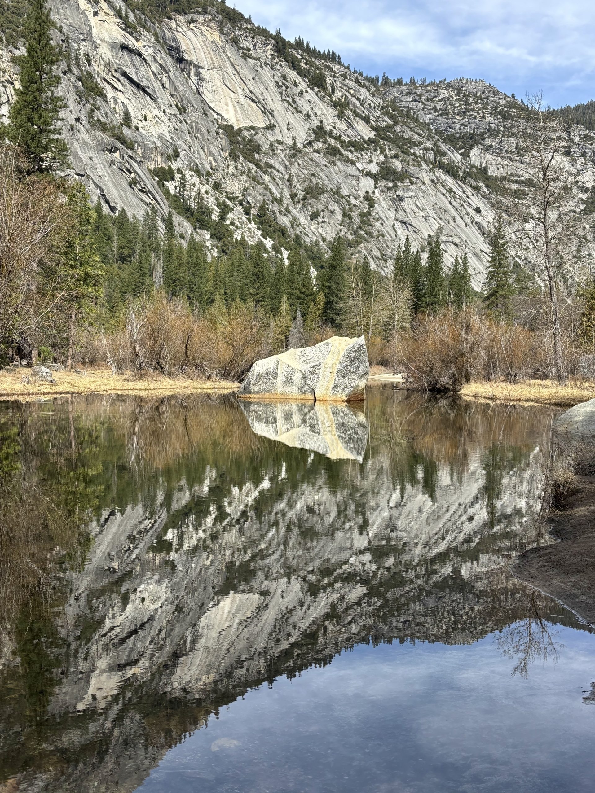 Granite cliff and pine forest reflected in a calm mountain pond, with a large boulder at the water’s edge.