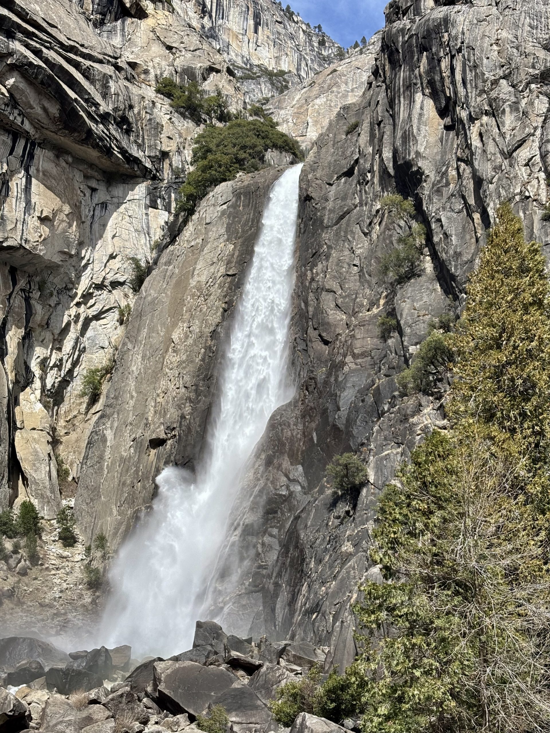 Tall waterfall cascading down a steep granite cliff with mist at the base and evergreen trees in the foreground.