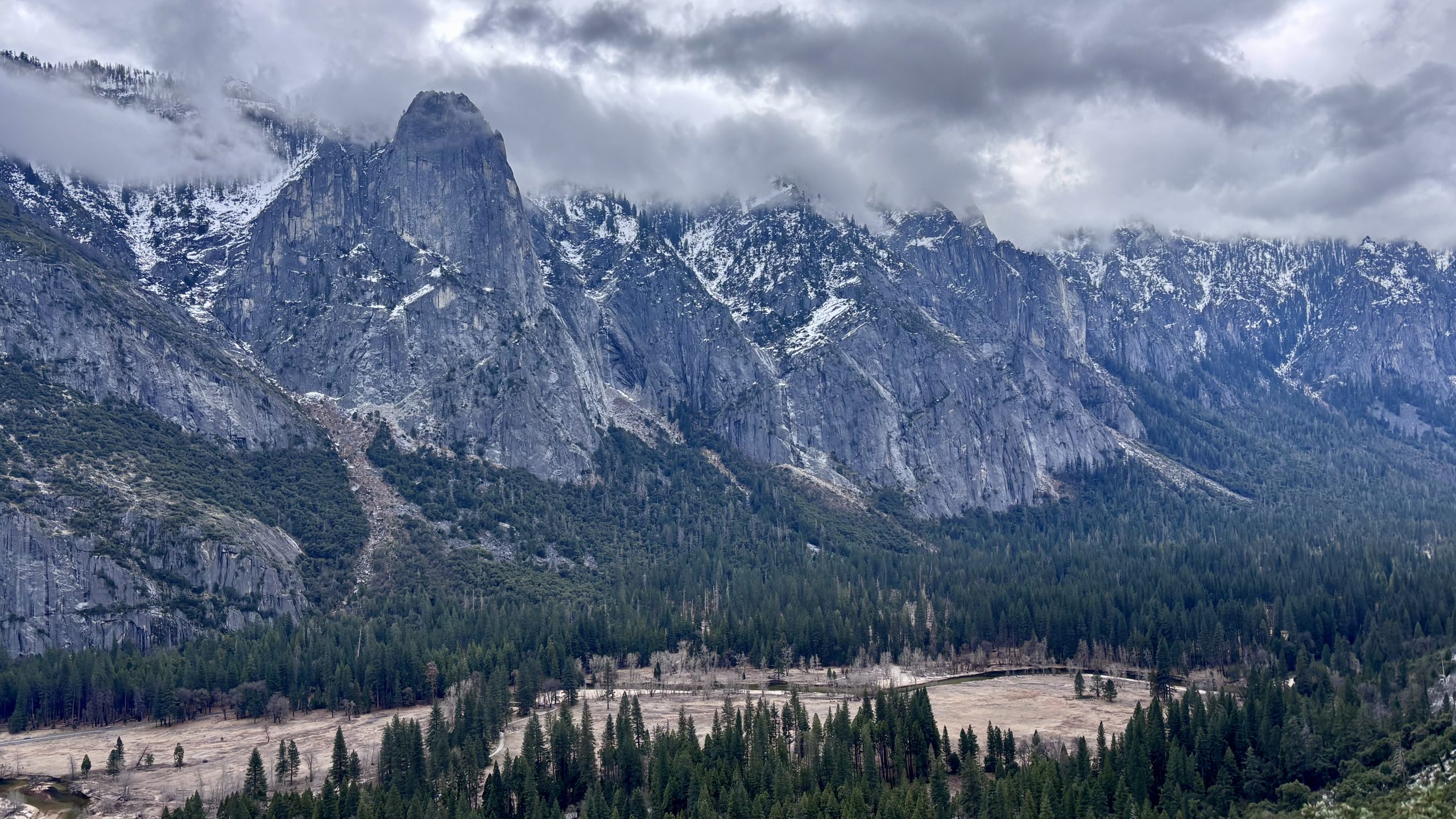 Panoramic view of a snow-dusted mountain valley with granite cliffs, forested floor, and cloudy sky.