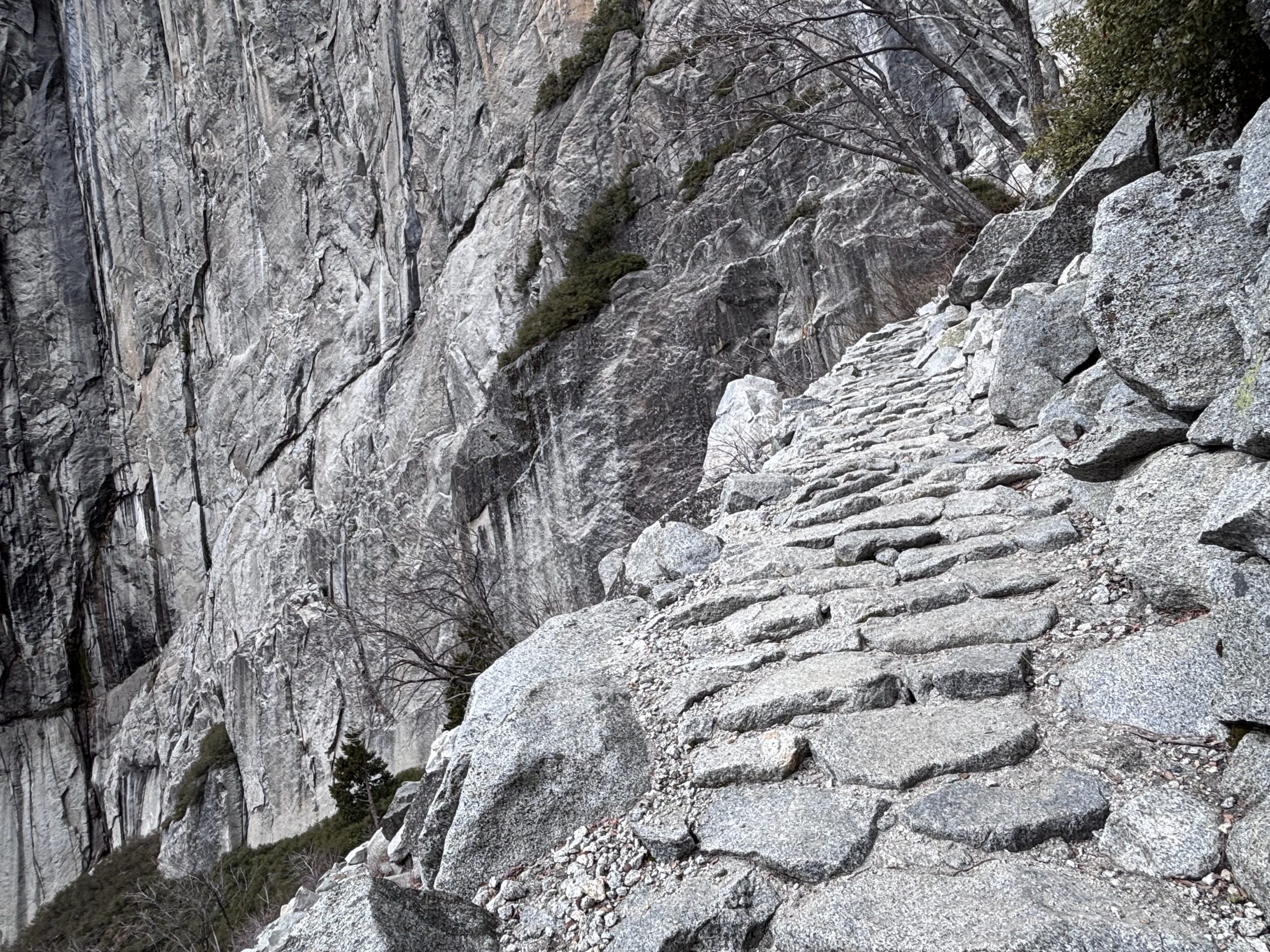 Stone steps carved into a steep mountain trail winding past a sheer granite cliff with sparse trees and boulders.