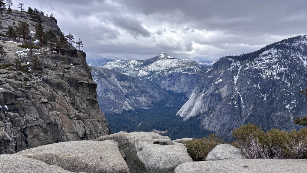 Snow-dusted granite cliffs and forested valley under a cloudy sky, viewed from a rocky overlook.