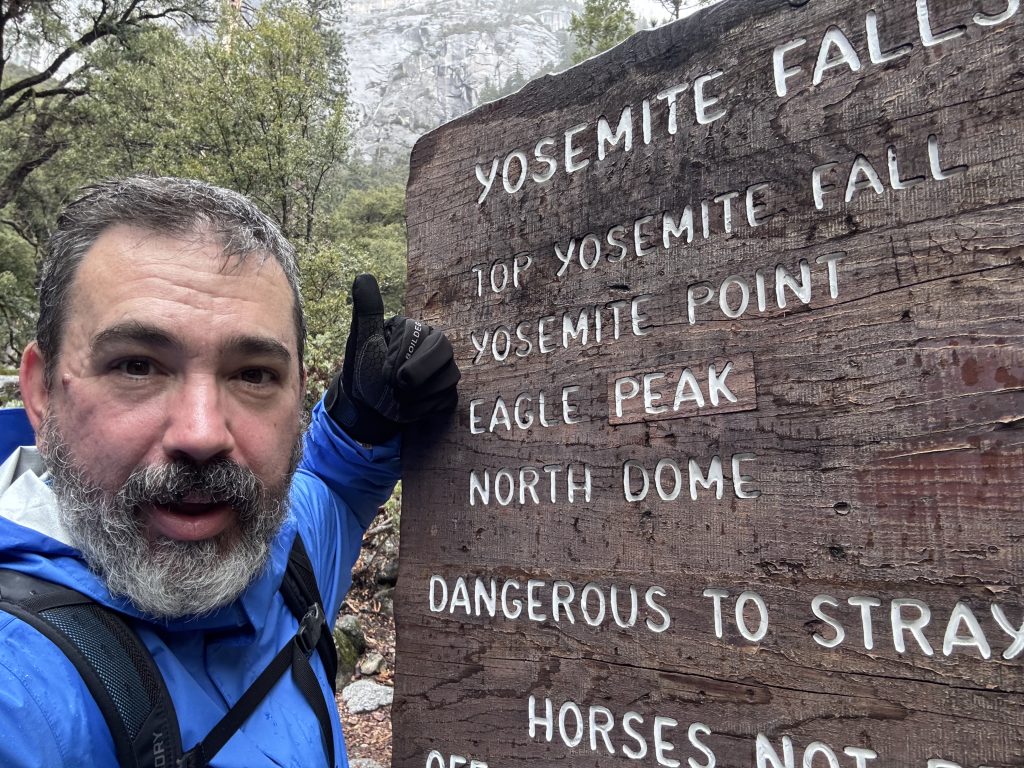 Man in hiking gear giving a thumbs-up next to a wooden Yosemite trail sign listing Yosemite Falls, Yosemite Point, Eagle Peak, and North Dome.