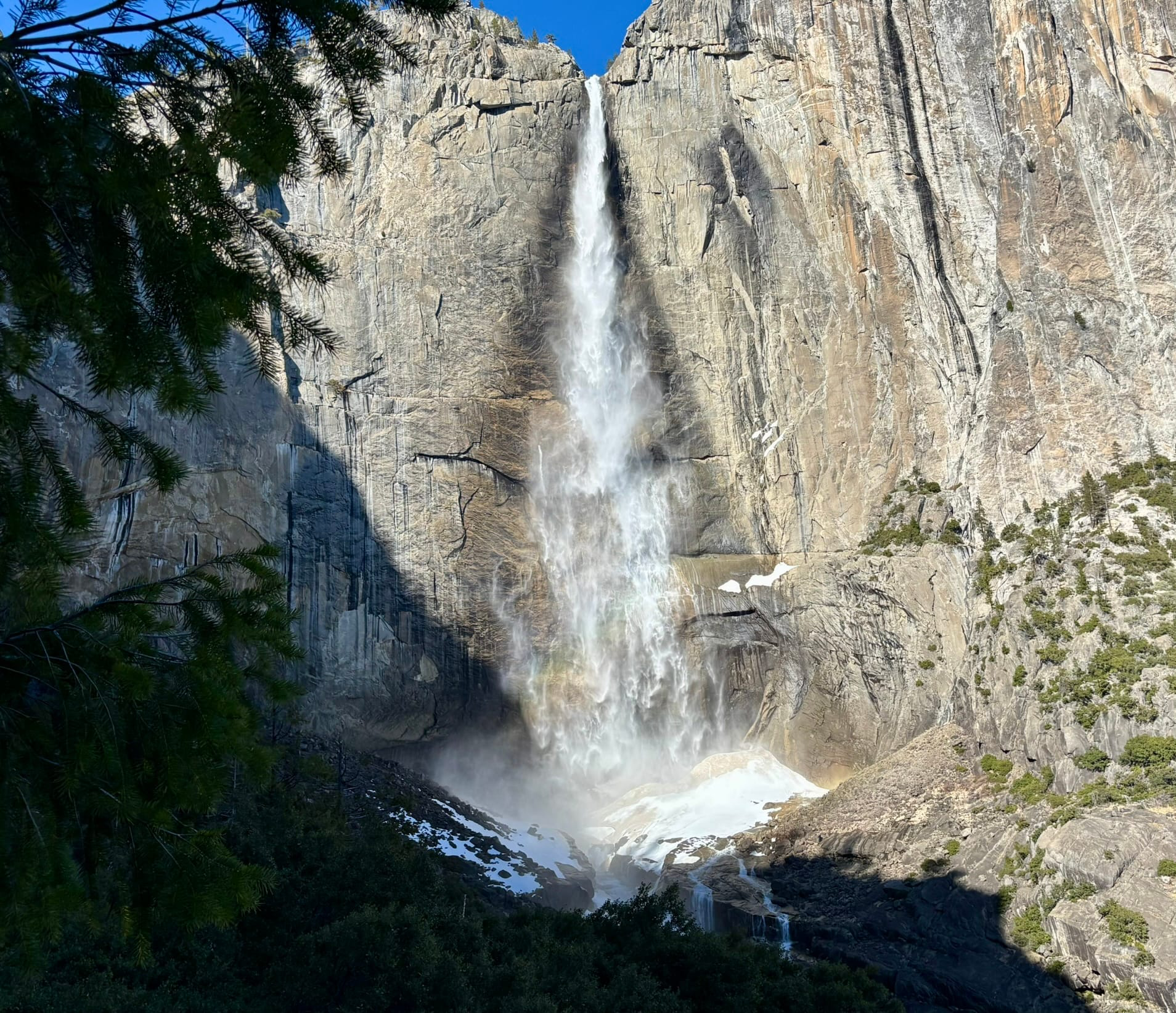 Tall waterfall plunging down a sheer granite cliff with spray and a faint rainbow, framed by evergreen branches and rocky foothills with patches of snow.