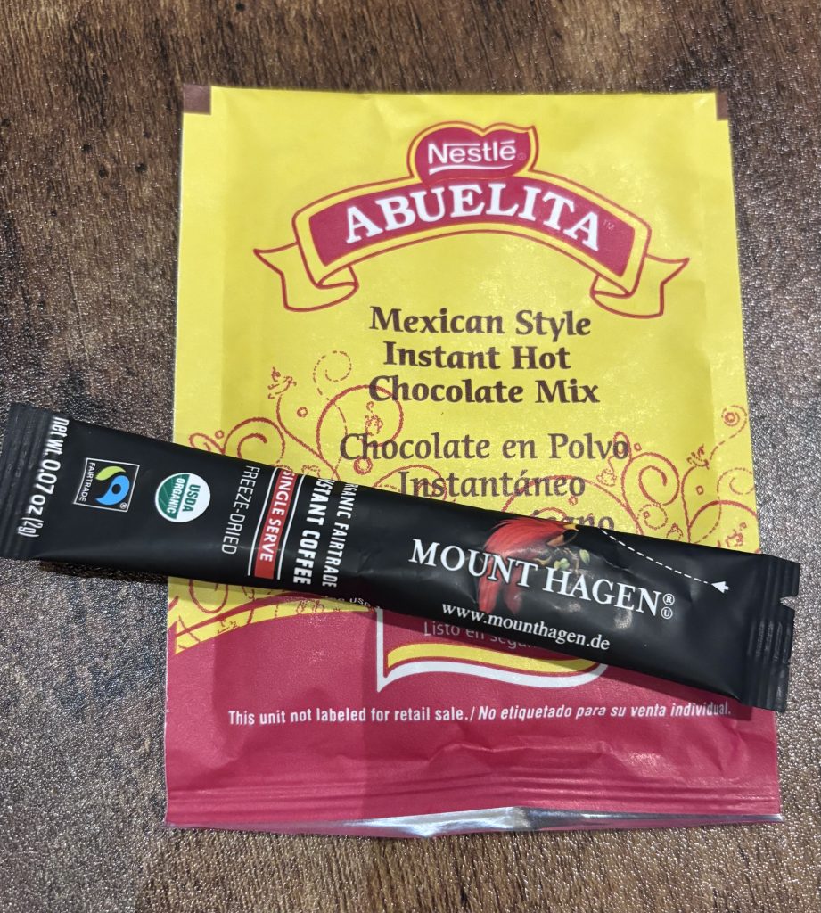 Yellow Nestlé Abuelita Mexican-style instant hot chocolate packet with a black Mount Hagen single-serve instant coffee stick placed diagonally across it.