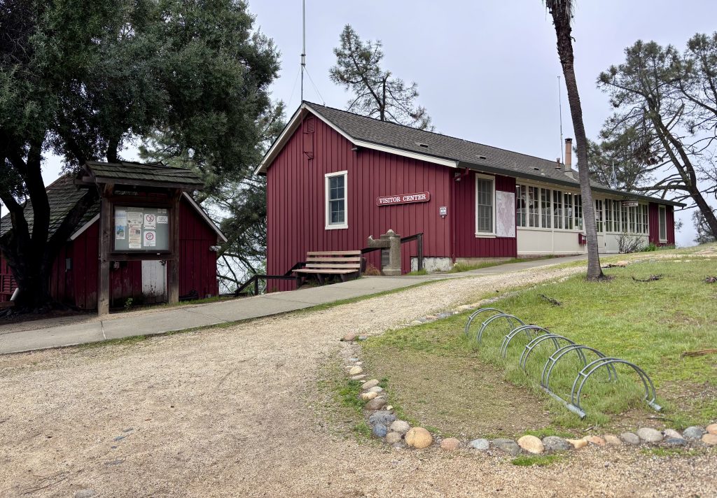 Red visitor center building painted deep red with a small notice board, bench, bike rack, trees, and gravel path in a park-like setting.