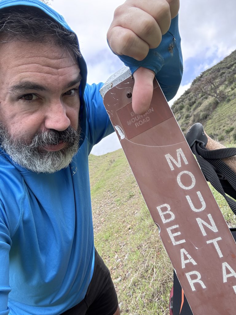 Bearded man in a blue hoodie giving a thumbs-down next to a worn trail sign that reads "Mount Bear/ Mountain Road" on a grassy hillside.