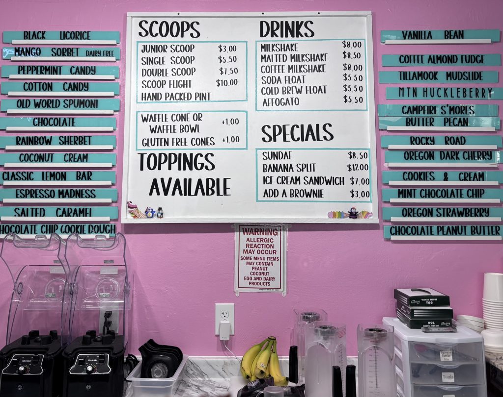 Colorful ice cream shop menu on a pink wall, listing scoop sizes, drink options, and over 25 unique flavors like Black Licorice, Mountain Huckleberry, and Oregon Strawberry, along with toppings and dessert specials.