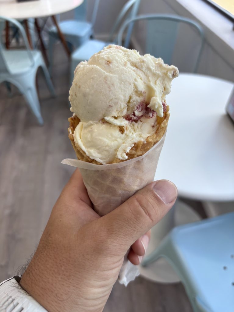 A hand holding a double scoop of ice cream in a waffle cone, wrapped in wax paper. The ice cream appears to be vanilla with swirls of fruit, possibly strawberry or raspberry. The background shows a light and airy ice cream shop interior with pale blue metal chairs and round white tables.