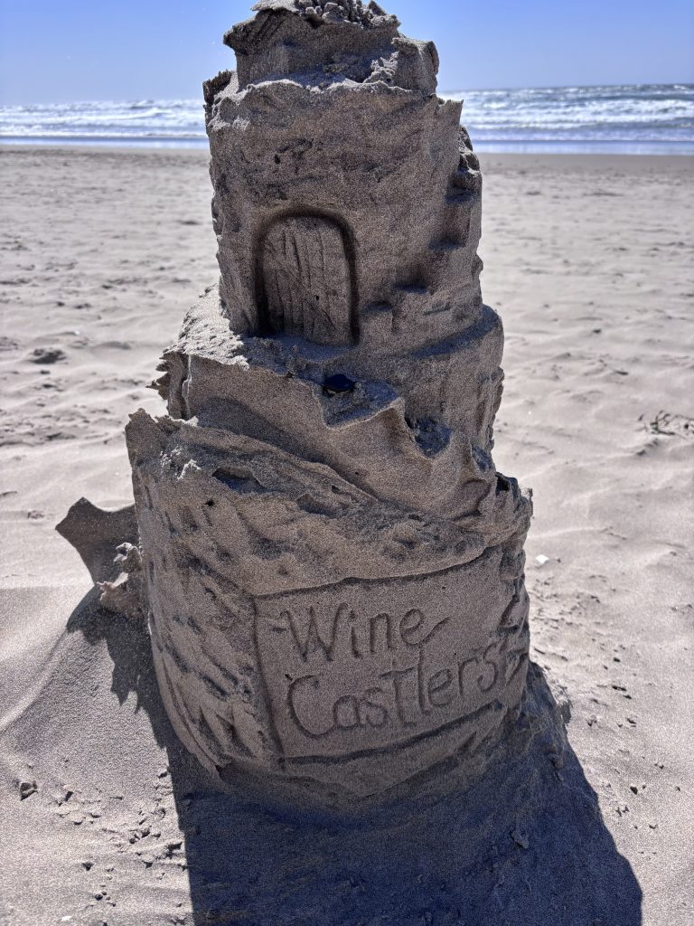 A close-up of a tall sandcastle on a beach with “Wine Castles” carved into the base. The detailed structure features stacked tiers, a carved door, and textured walls, with the ocean and horizon in the background under a clear sky.