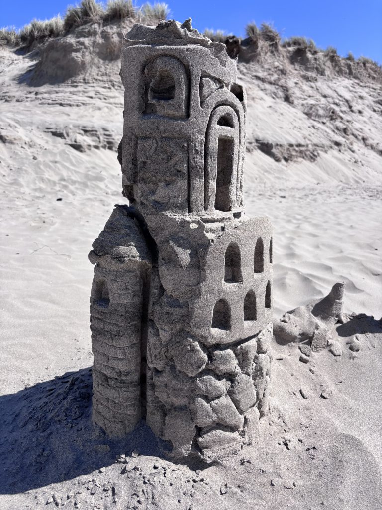 An intricately sculpted sandcastle with a cylindrical tower and arched windows, standing on a dry, sandy beach with dune grass and a blue sky in the background.