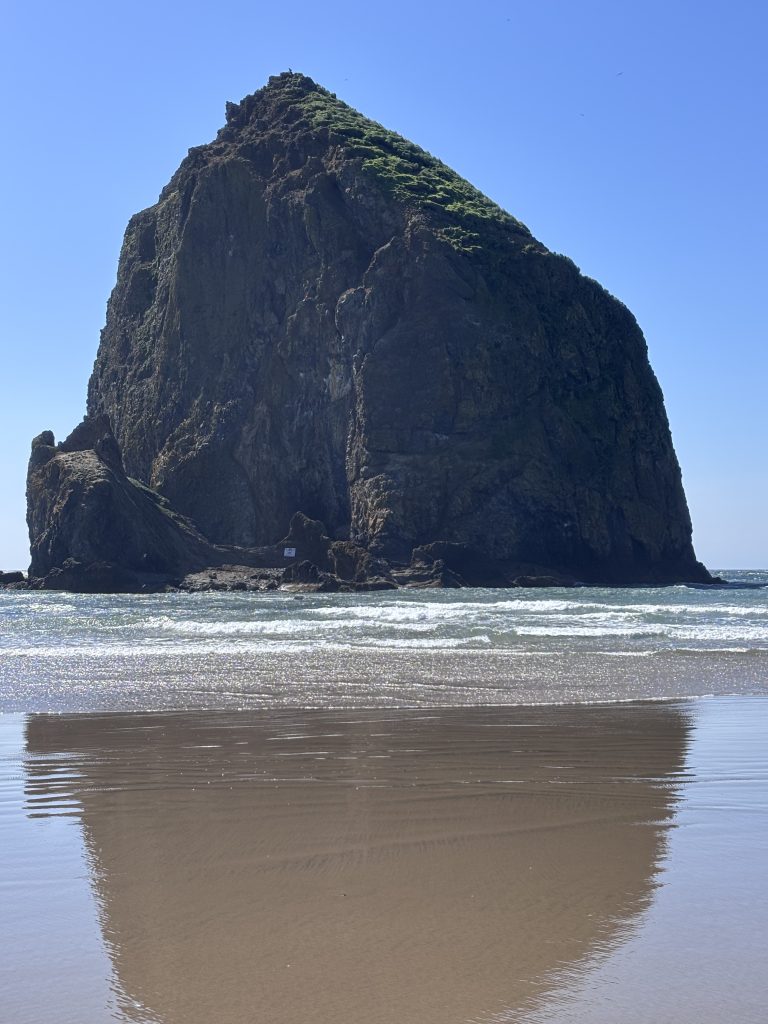 A towering view of Haystack Rock rising from the Pacific Ocean, with its green-topped peak reflecting in the wet sand at low tide.