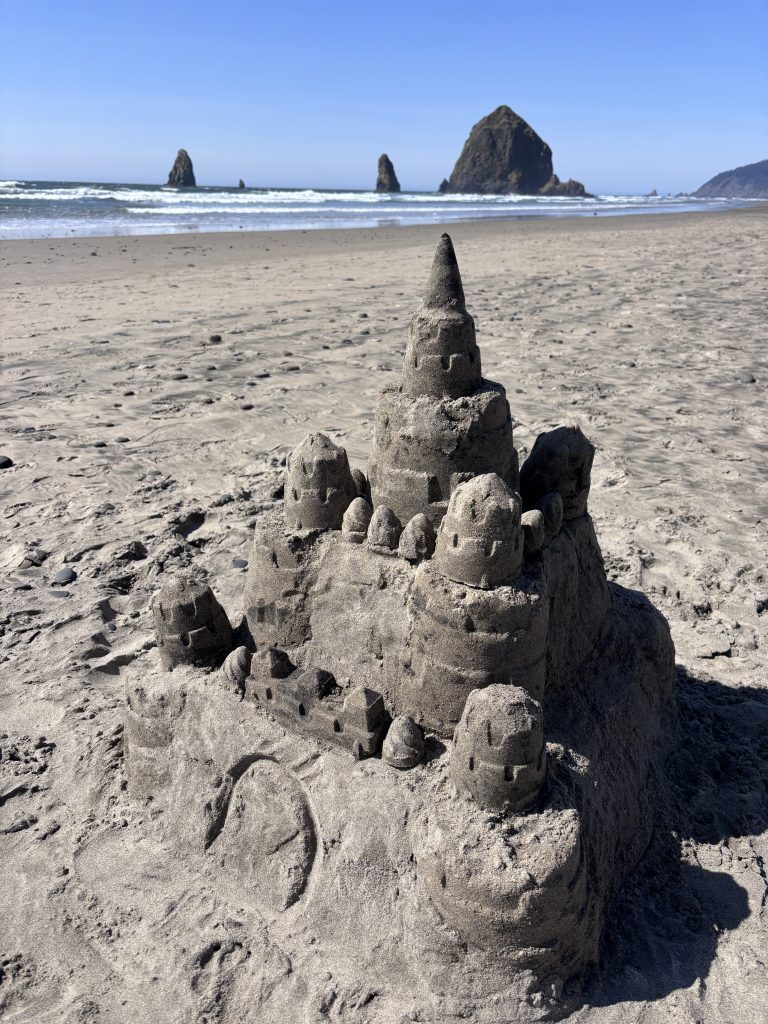 A classic sandcastle with multiple towers and a central spire, sitting on a flat beach with Haystack Rock and other sea stacks rising from the ocean in the distance.