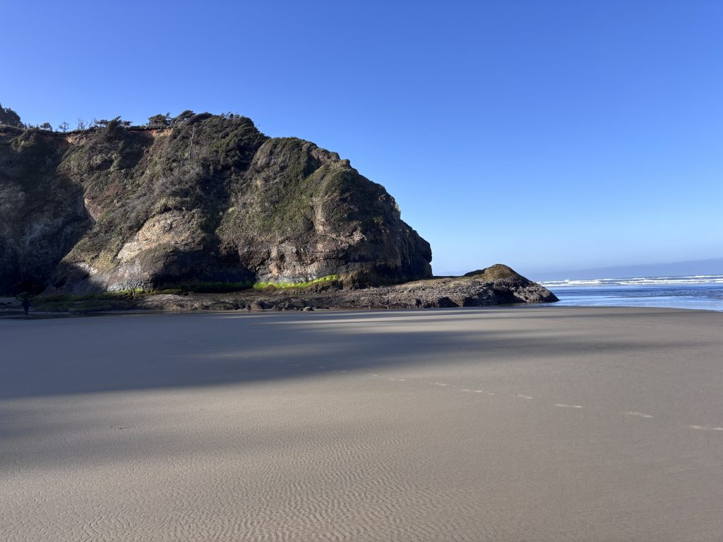 A wide-angle view of a sandy beach with a large, moss-covered rock formation dominating the left side of the frame. The ocean stretches out to the horizon under a clear blue sky. Light and shadow play across the sand, creating a textured effect.