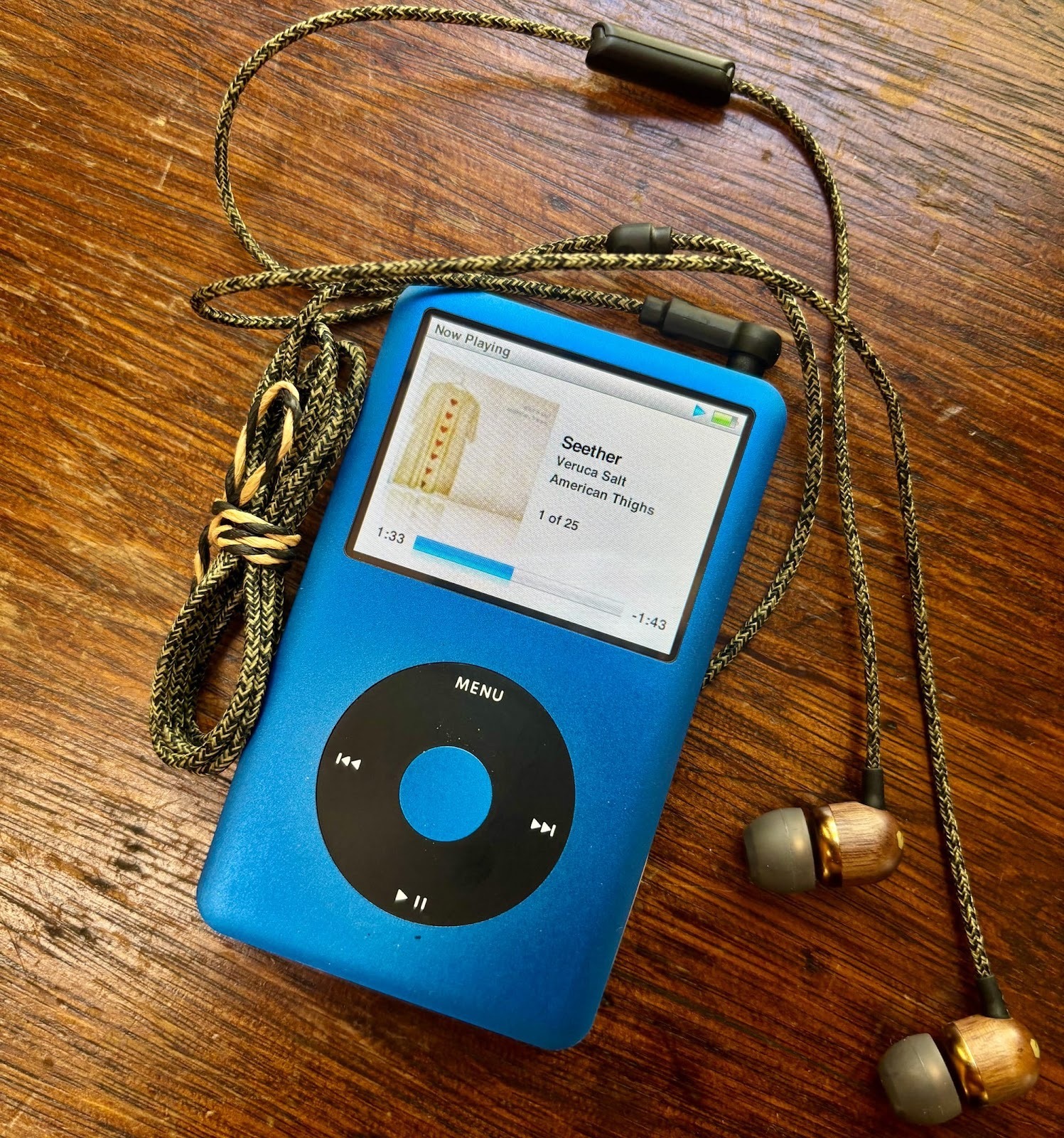 Blue iPod Classic playing the song “Seether” by Veruca Salt from the album American Thighs, with braided wired earphones resting on a wooden table.