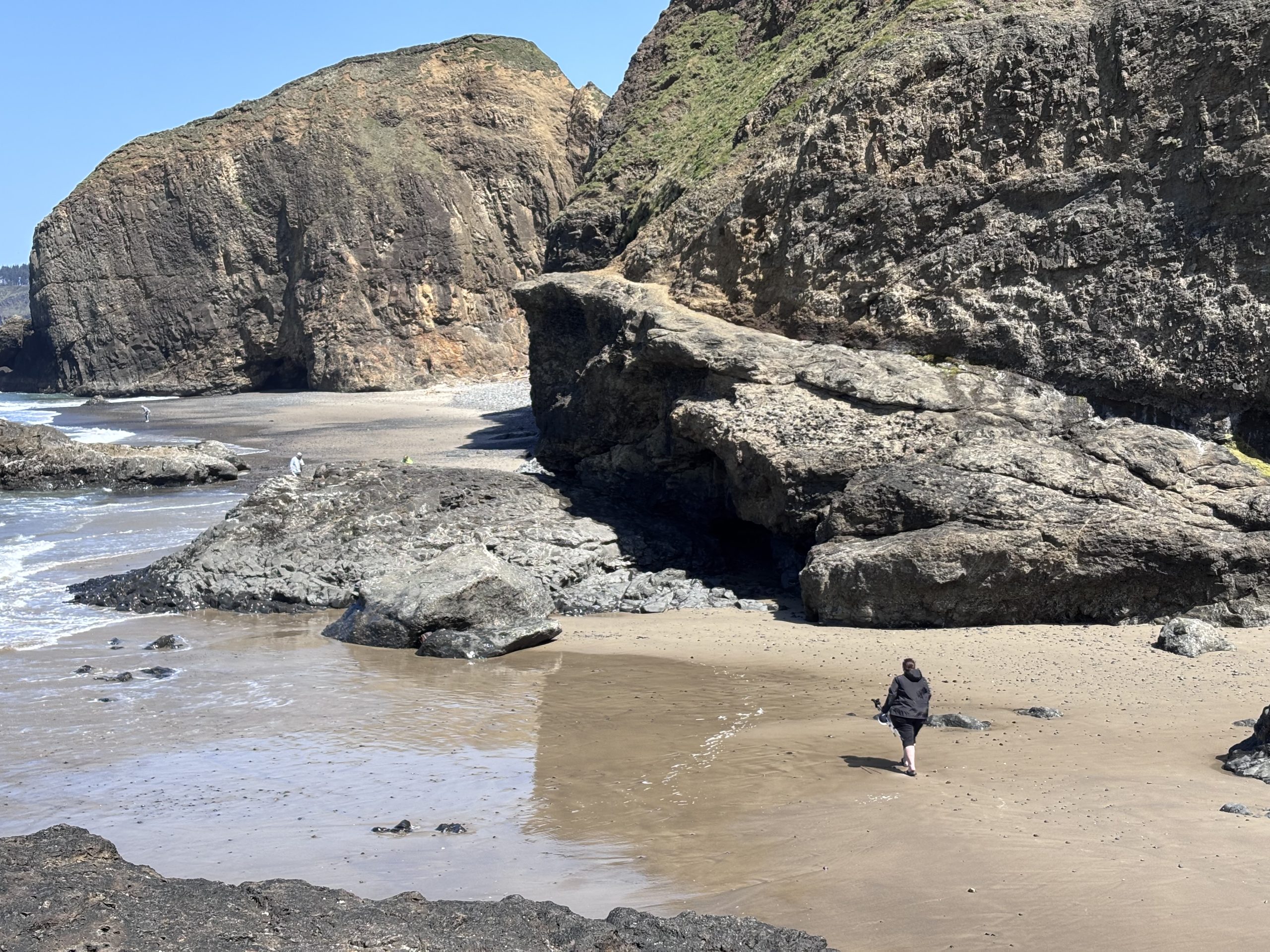 Person walking barefoot across a sandy beach surrounded by large coastal cliffs and sea stacks on a sunny day along the Oregon Coast.