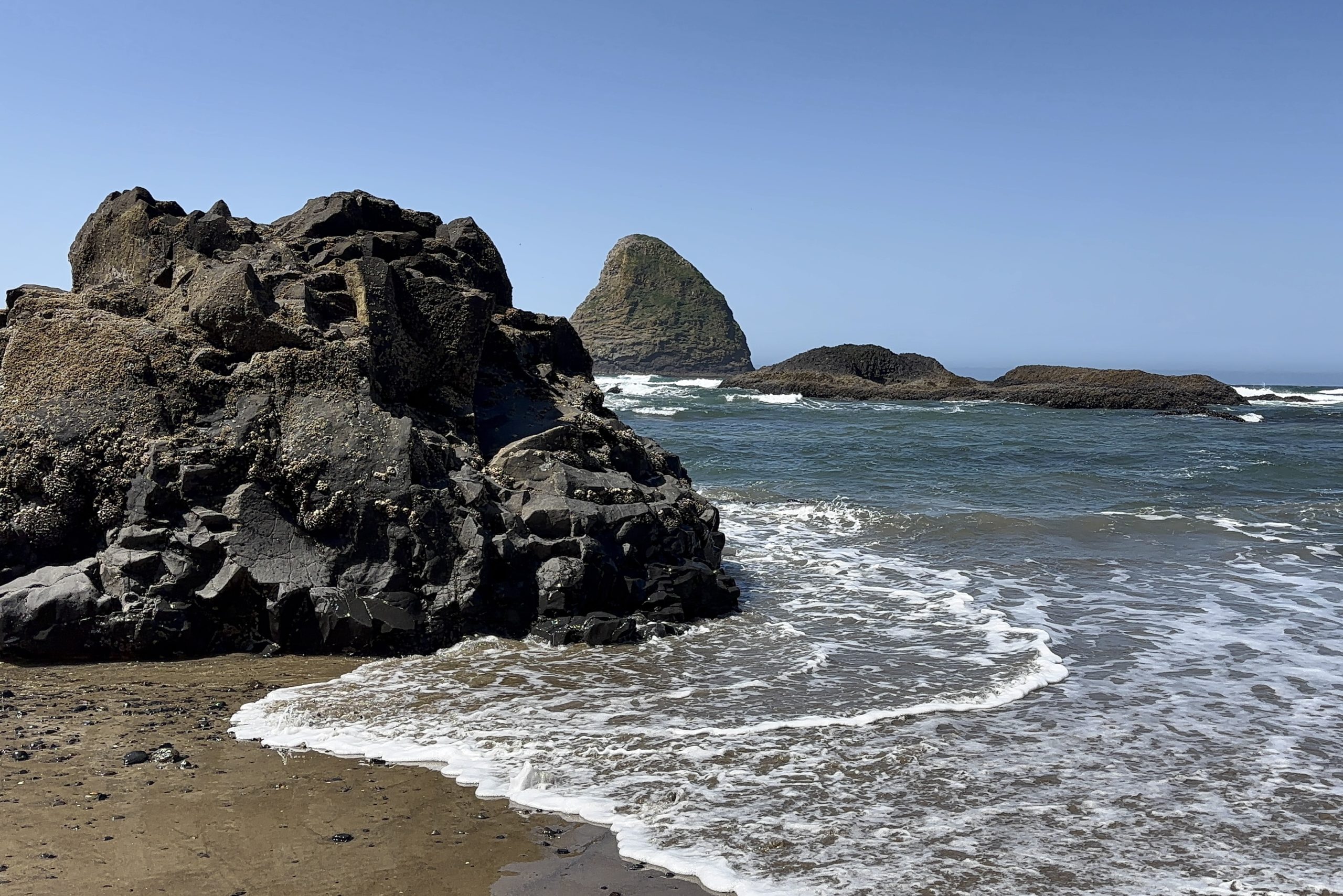 Rocky sea stack rising from the ocean near the shore, with waves gently washing against dark basalt formations under a clear blue sky along the Oregon Coast.