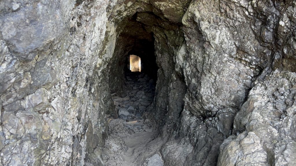 Narrow sea cave tunnel carved through rugged coastal rock, with a view of daylight and the beach visible through the far opening.