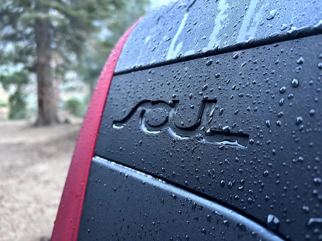 A close-up shot of the rear Kia Soul badge with water droplets on the surface, framed by red taillight and blurred forest in the background.