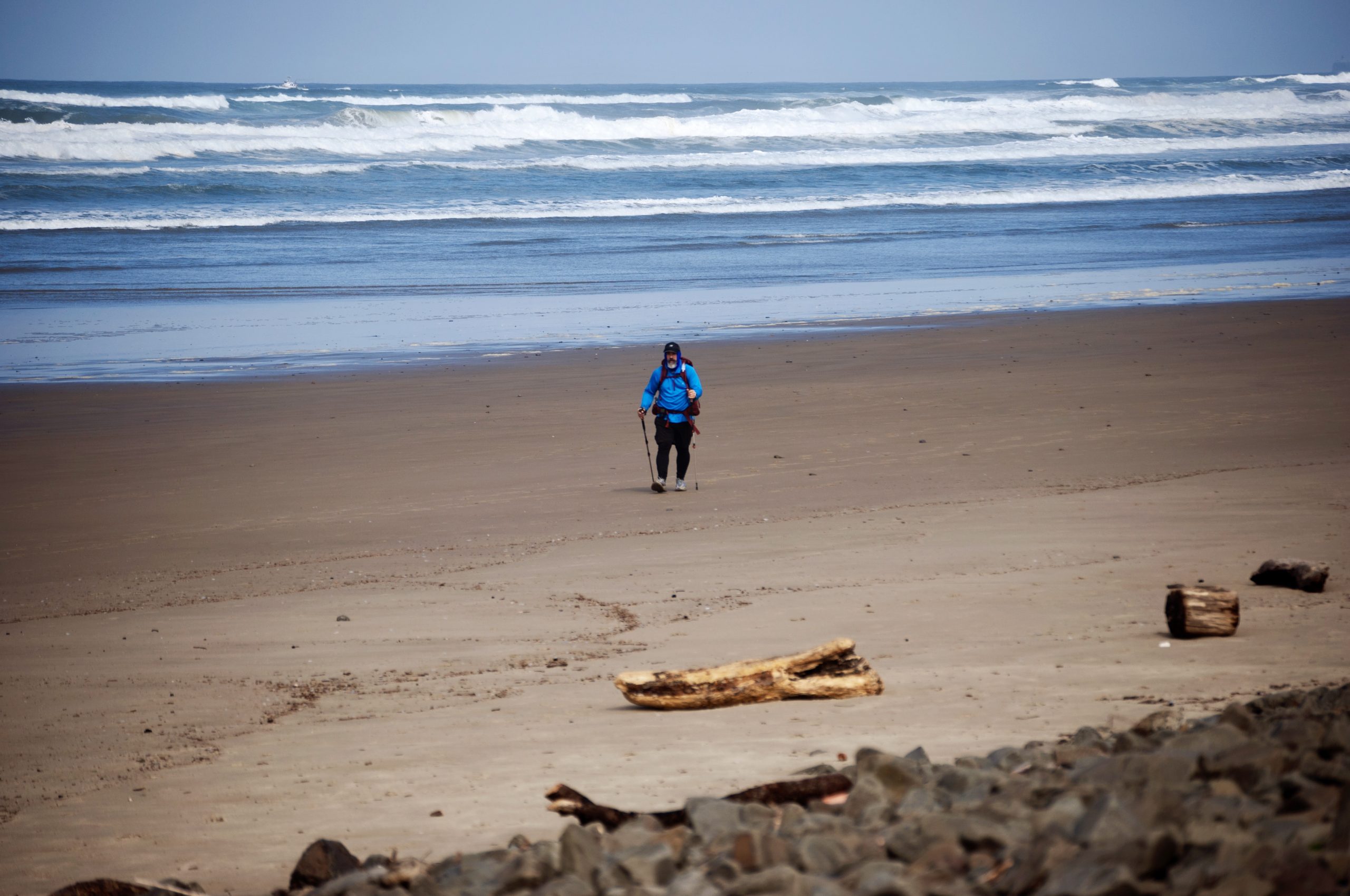 A lone figure is walking along a vast, sandy beach towards the ocean. The person is wearing a bright blue jacket and is using walking sticks. The sky is overcast and the water stretches out to the horizon.