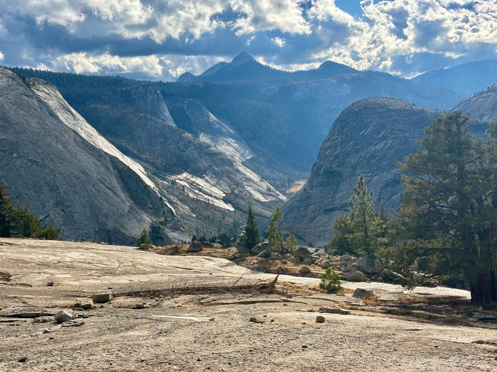 A wide, sunlit view of a granite valley with towering cliffs and coniferous trees under a partly cloudy sky. The valley floor is covered in light-colored rock and gravel, and the cliffs rise sharply on either side.