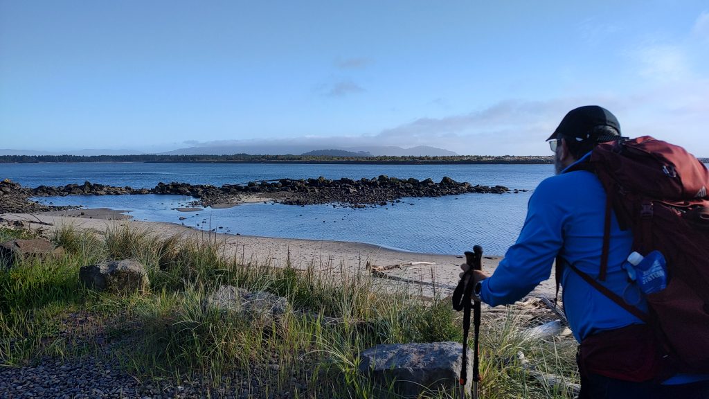 A person stands overlooking a view of a calm body of water with rocky outcroppings visible at low tide. They appear to be hiking, with a backpack and hiking poles visible.