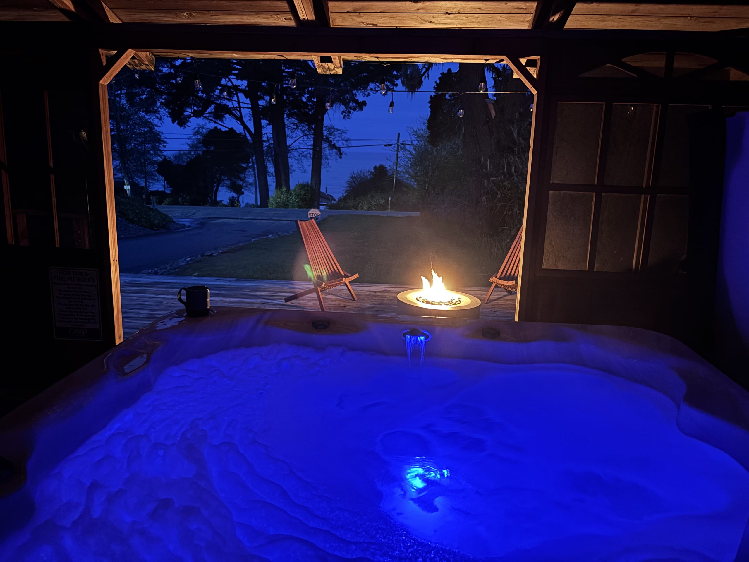 Private hot tub with blue LED lighting inside a cozy wooden cabin, overlooking a fire pit and two Adirondack chairs at twilight. Ocean and trees visible in the background