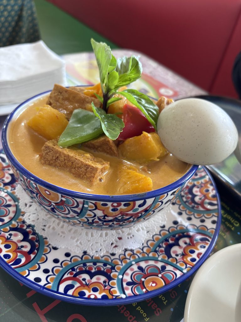Colorful ceramic bowl of Thai pineapple curry with tofu, red bell pepper, snap peas, and fresh basil, served with a hard-boiled egg on the side. Placed on a patterned doily and plate.