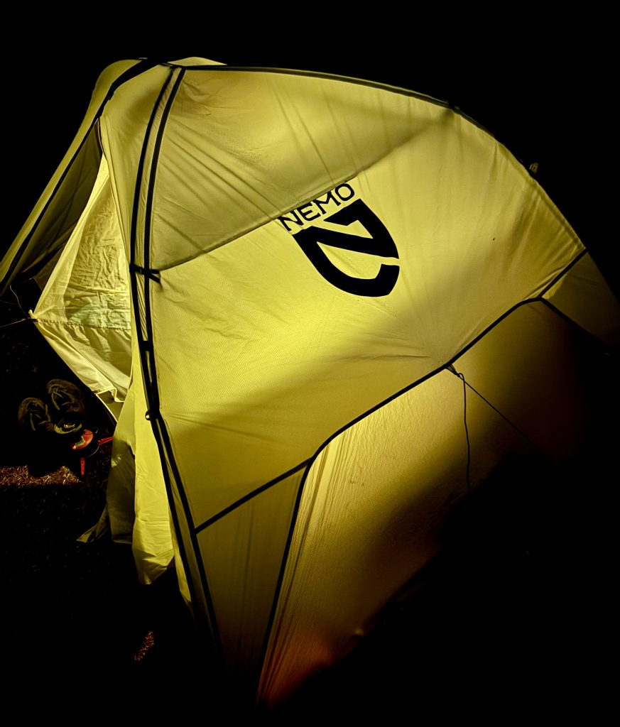 Glowing yellow Nemo-brand tent lit from the inside during nighttime camping, with hiking shoes and a fuel canister barely visible outside the entrance.