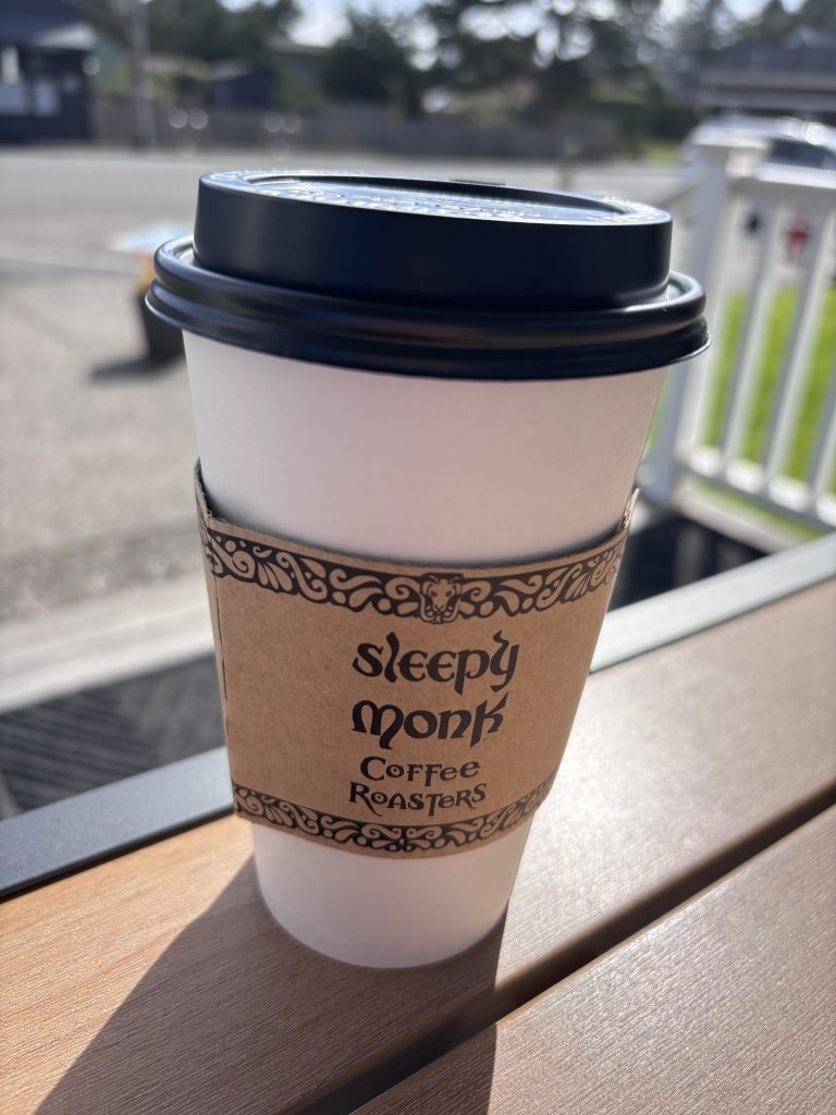 A to-go cup of coffee from Sleepy Monk Coffee Roasters sits on a wooden outdoor table, slightly steaming in the morning sun. The ornate sleeve and lid are in sharp focus, while the background blurs into a quiet neighborhood street