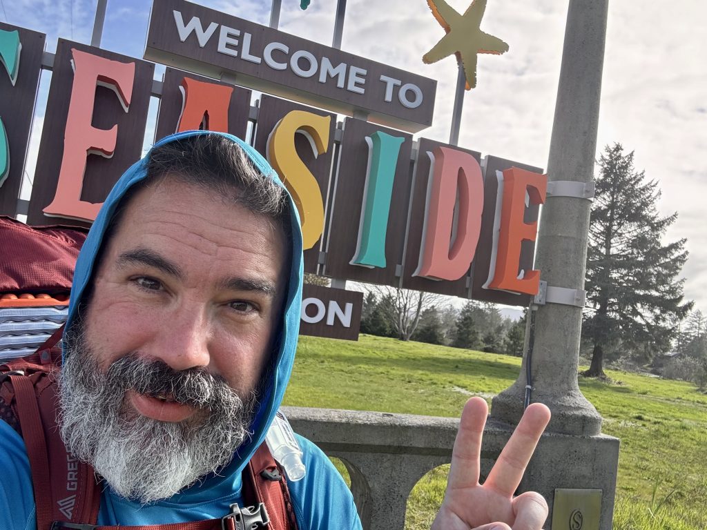 A smiling bearded hiker flashes a peace sign in front of the colorful “Welcome to Seaside” sign in Seaside, Oregon. He wears a blue hoodie and a red backpack with foam sleeping pad visible, under bright skies and evergreen trees.