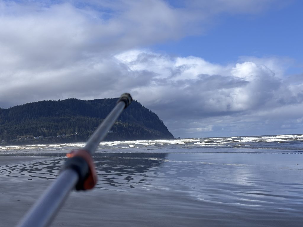Hiking pole pointing toward the misty, forested summit of Tillamook Head on a reflective beach at low tide along the Oregon Coast Trail.