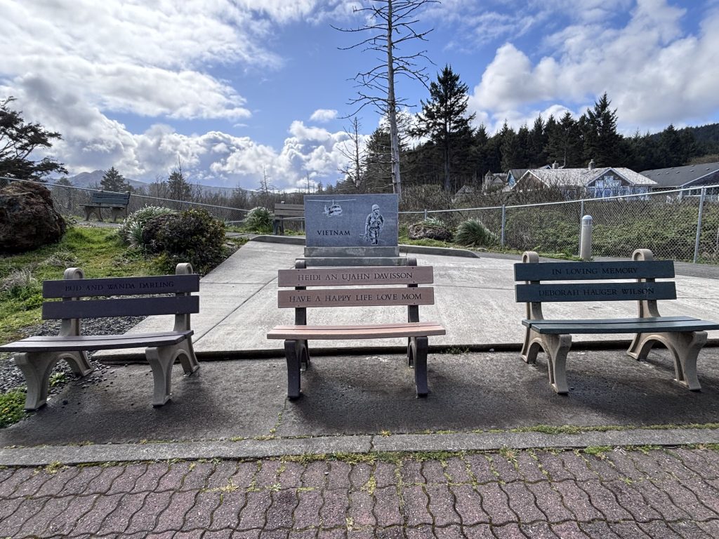 Vietnam memorial flanked by three personalized tribute benches overlooking a coastal town park in Oregon with cloudy skies and evergreen trees in the background.