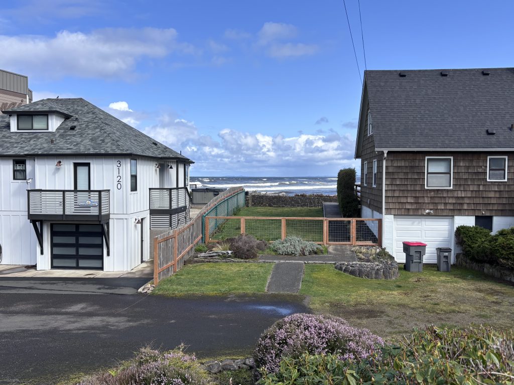 Ocean view framed between two beach houses in Seaside, Oregon, with a glimpse of crashing waves and coastal fencing leading to the water.